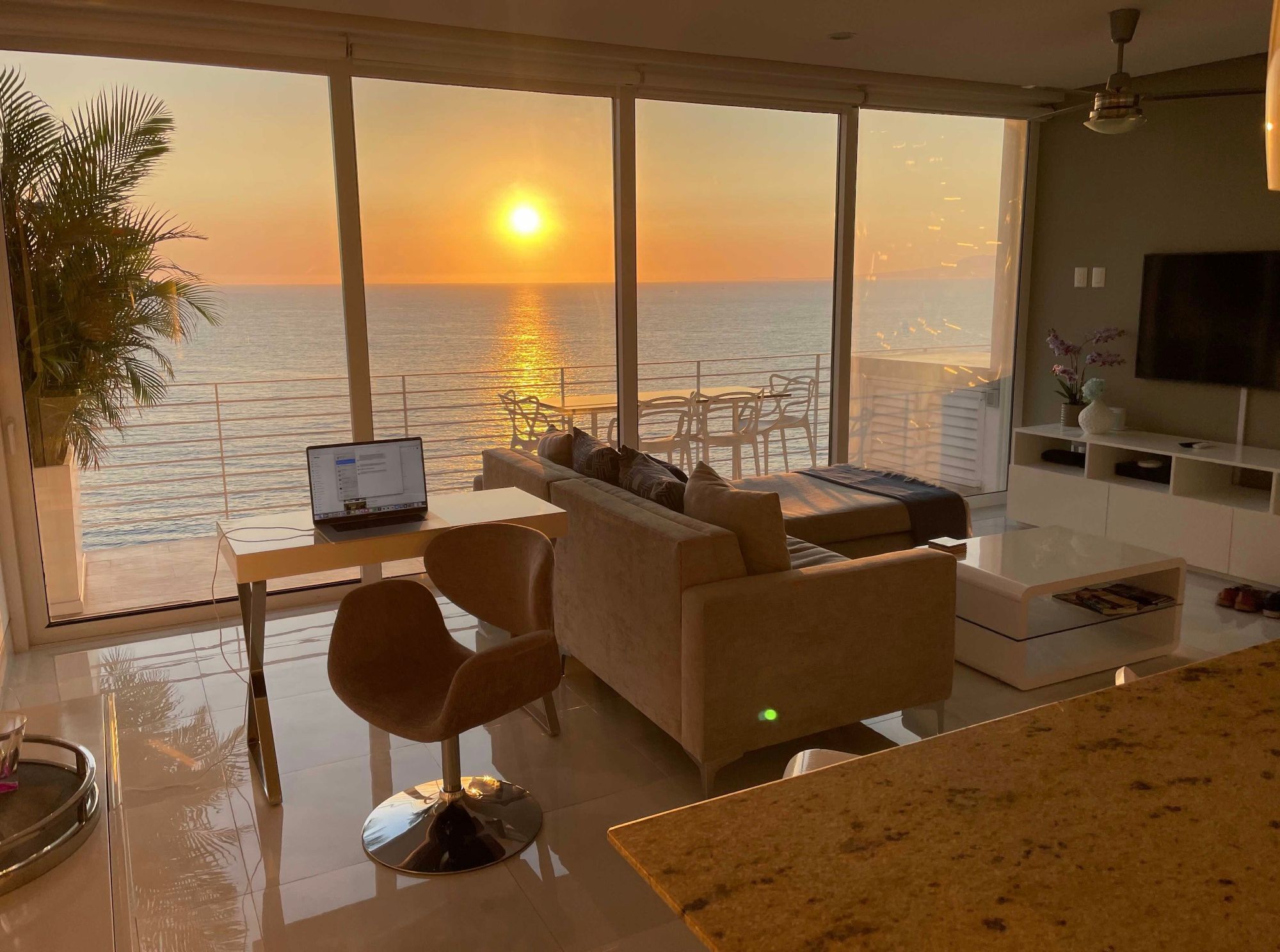 Photo shows a laptop set on a desk in front of a wall of windows, with a gorgeous sunset over the ocean in the distance. End description.